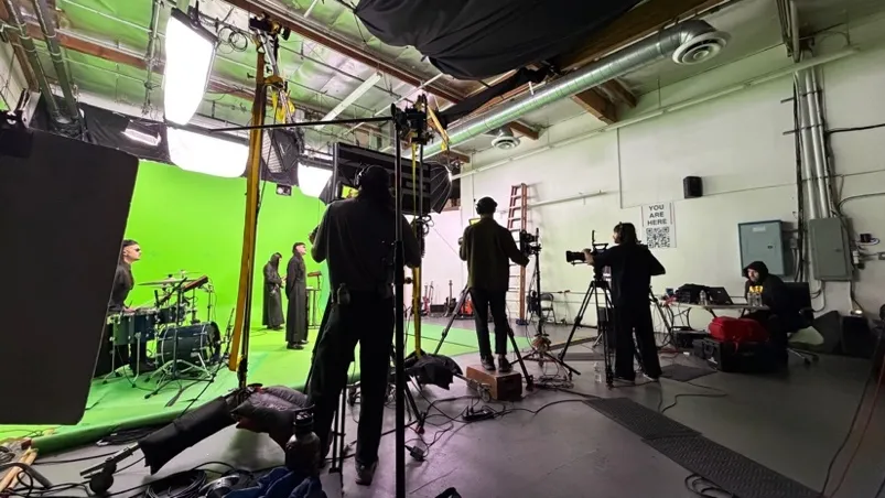 A sound stage during a shoot: a lit green screen, a drum kit, and three camera operators working behind their tripods.