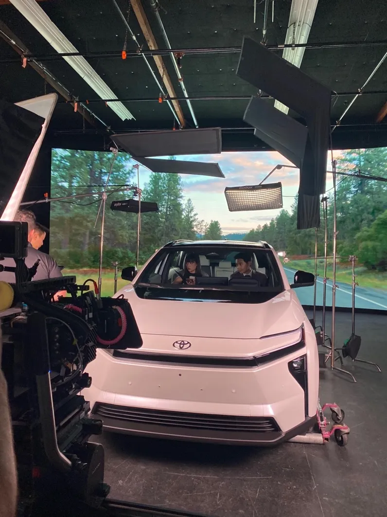 A car on a sound stage in front of a large LED wall showing a forest road, ringed by lighting flags and stands, with a cinema camera in the foreground.